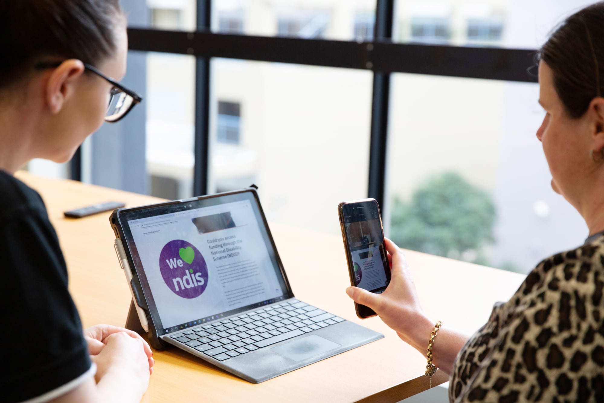 Two people looking at a laptop screen. One is holding a phone to the laptop screen and the other is watching on.