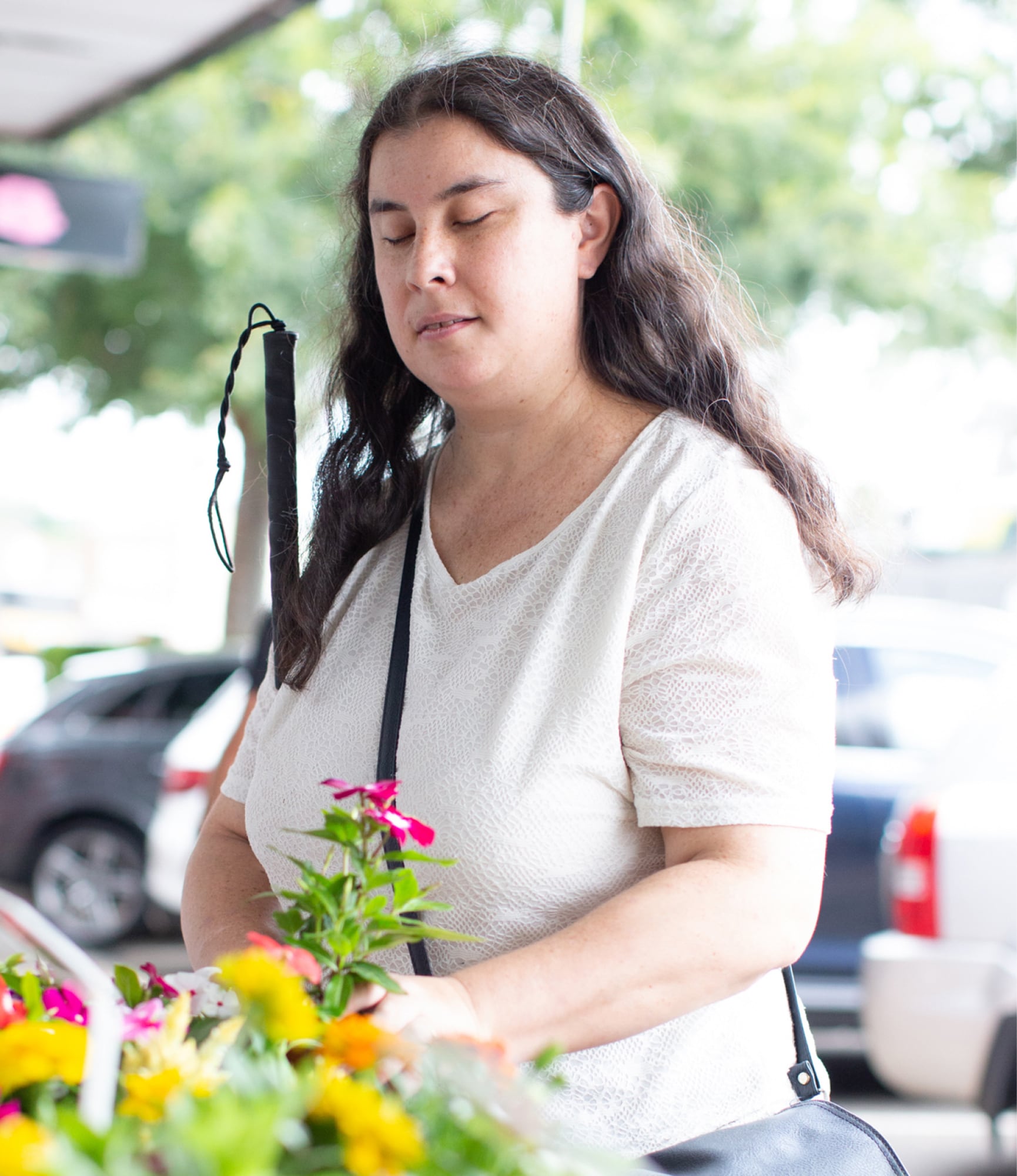 A person is standing in the street holding some flowers in one hand and their white cane in their other arm.