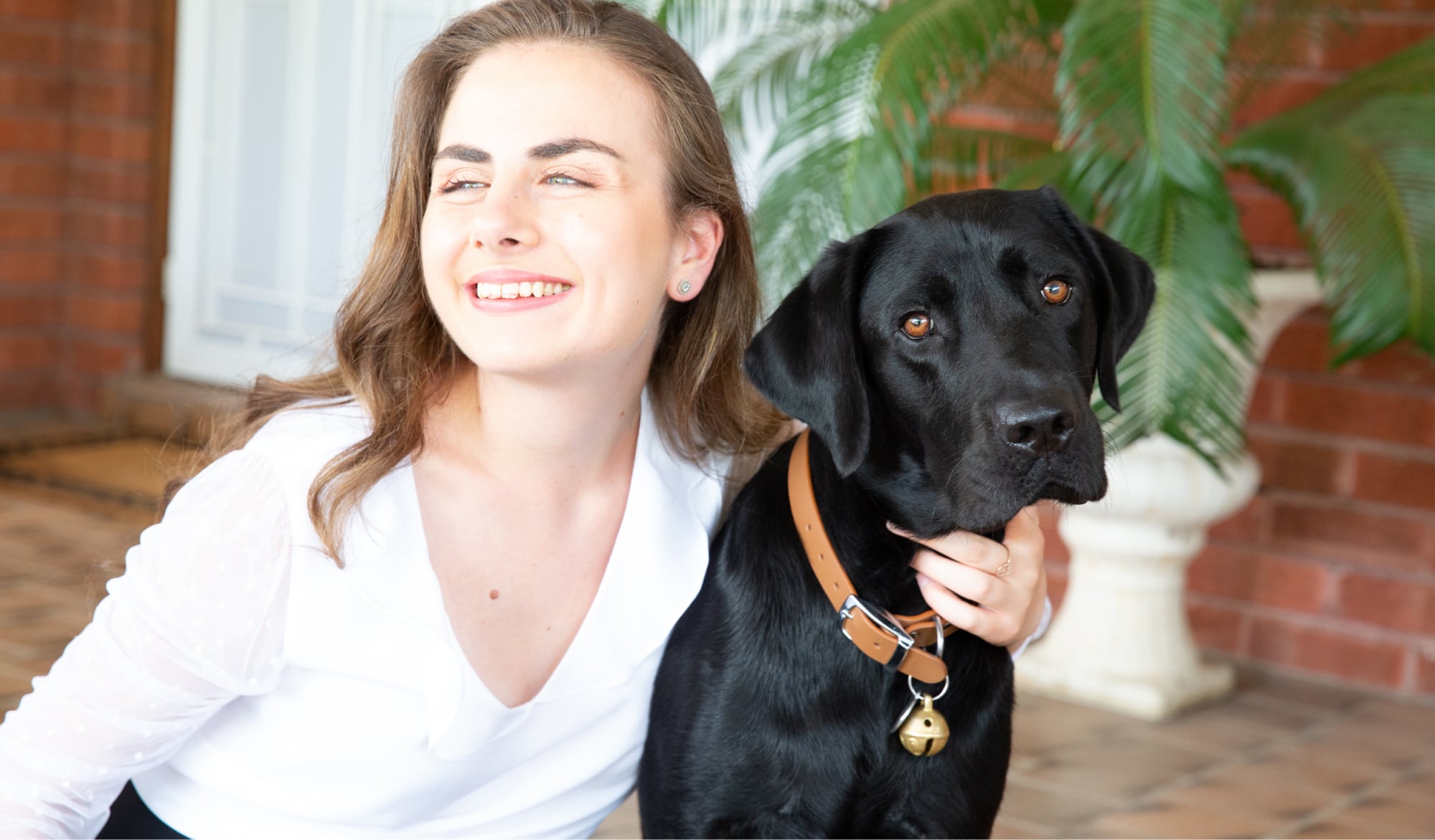 A person looking to the left of the camera smiling with their arm around the neck of a black labrador dog.