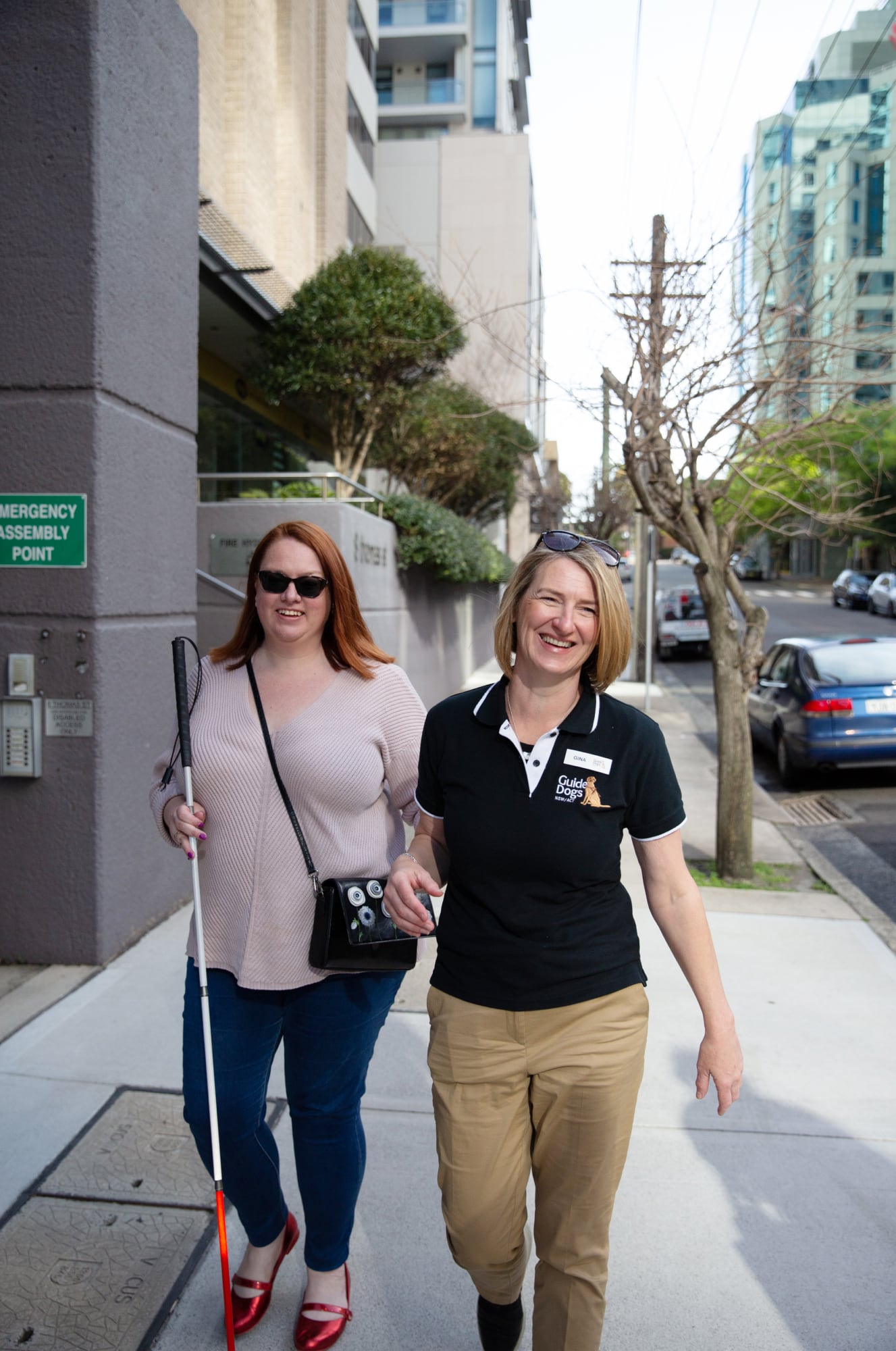 A person with their white cane walking down the street with a Guide Dogs staff member. Both people are smiling at the camera and the person with the cane is holding the staff member by the elbow.