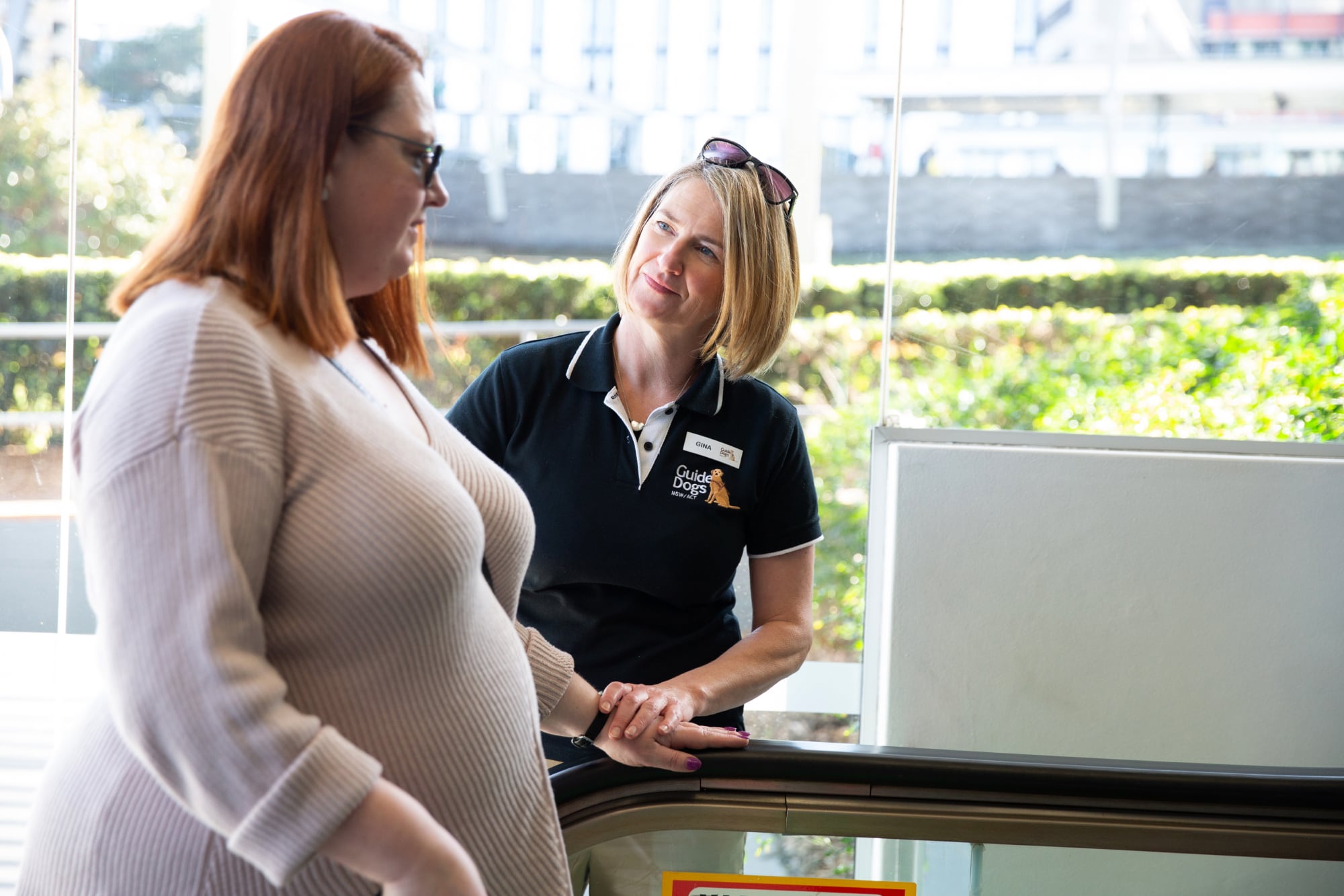 A Guide Dogs staff member with a person who is getting onto an escalator. The staff member is looking at the person.