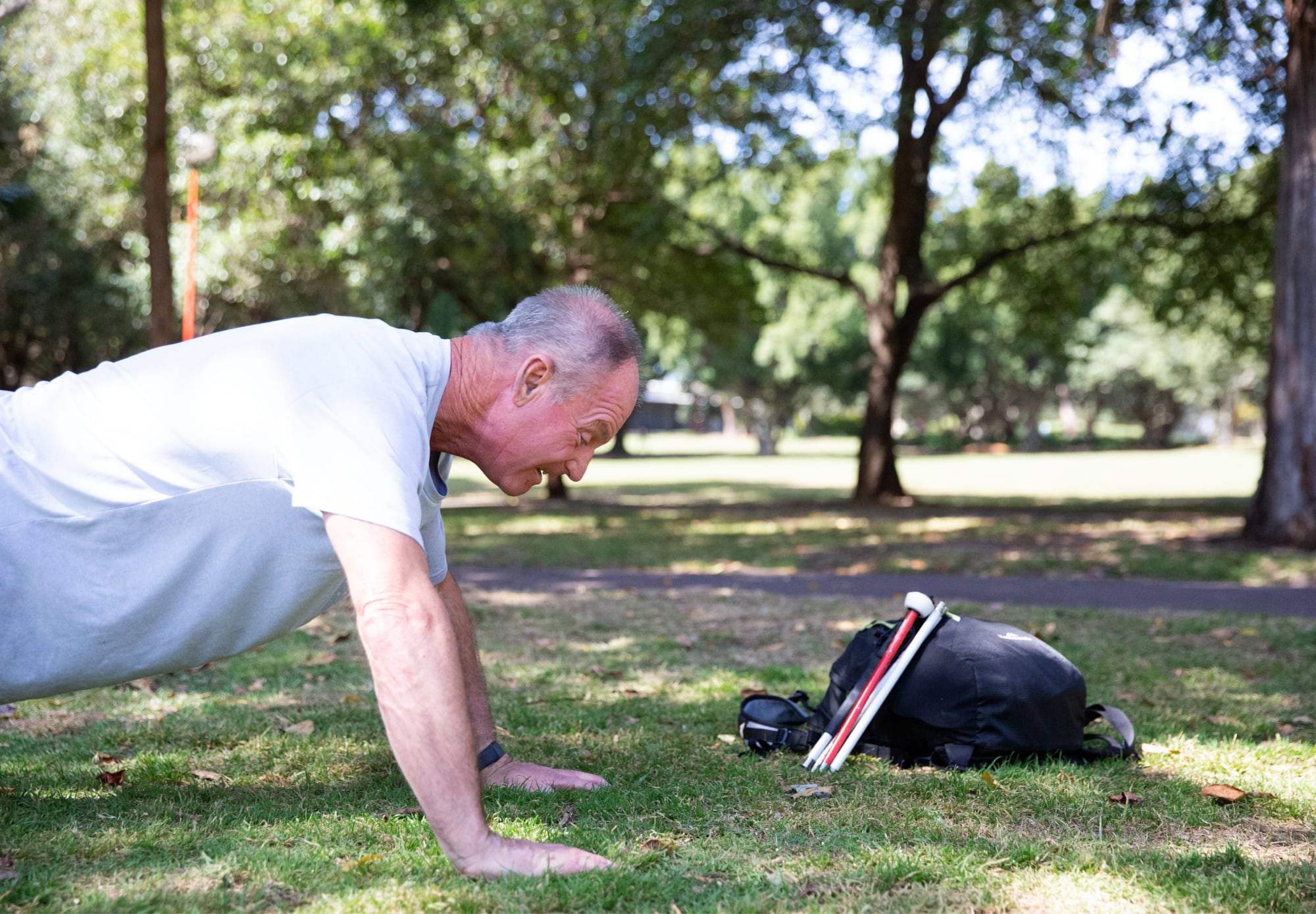 A person doing a pushup on grass, with their cane and a backpack beside them.