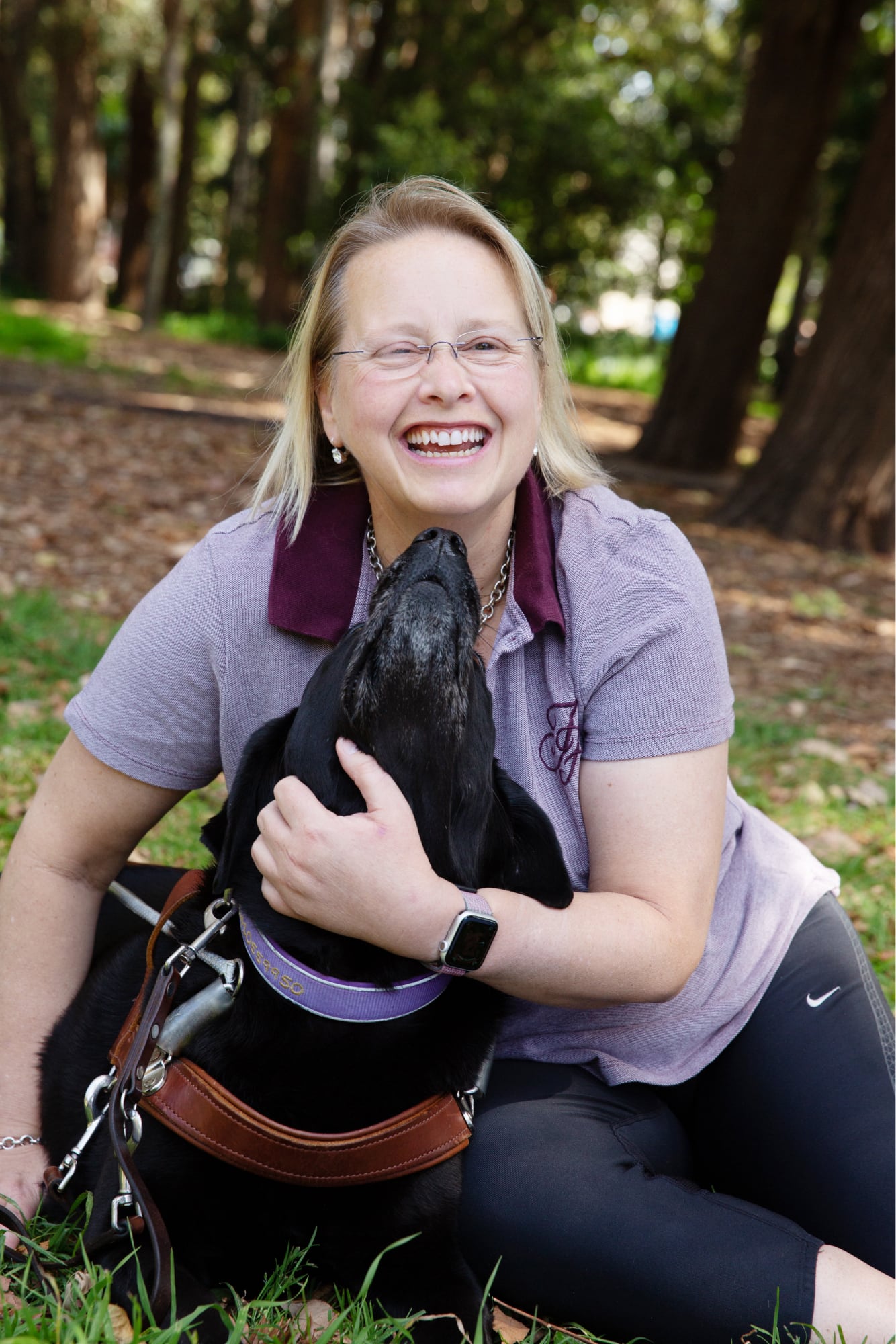 A person and their black labrador Guide Dog outside in the park. The dog is looking up at the person and the person is smiling at the camera.