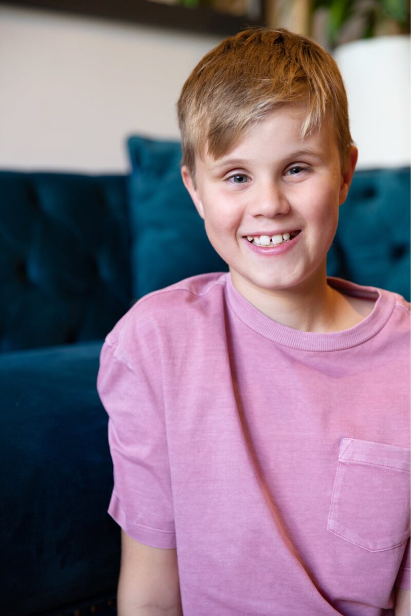 Close up of a young child with a big smile on their face. They are seated on the ground with a couch in the background.