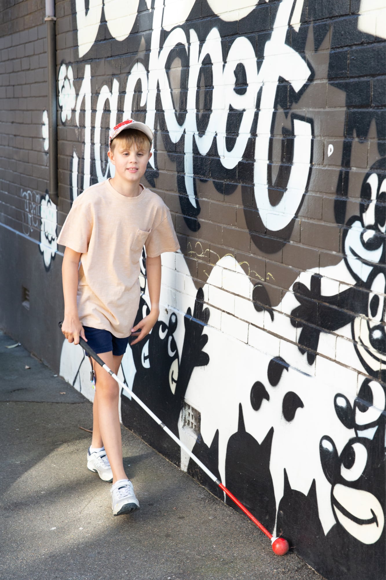 A child walking down the street using their white cane. The child is walking against a wall that has graffiti on it.