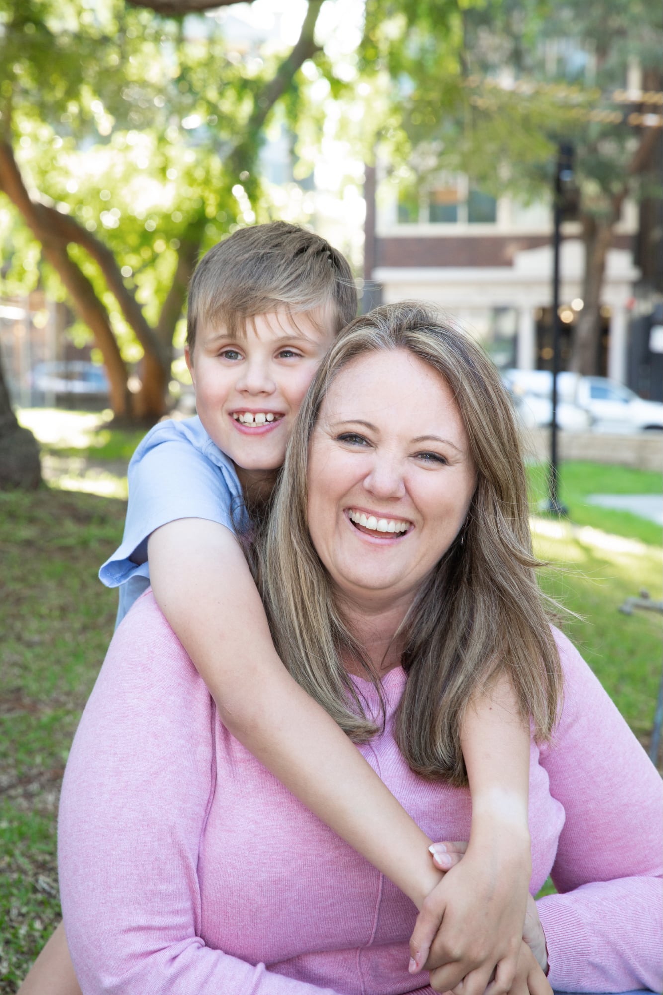Young child with their arms around their mothers neck and they are both looking at the camera smiling. They are seated on grass outside.