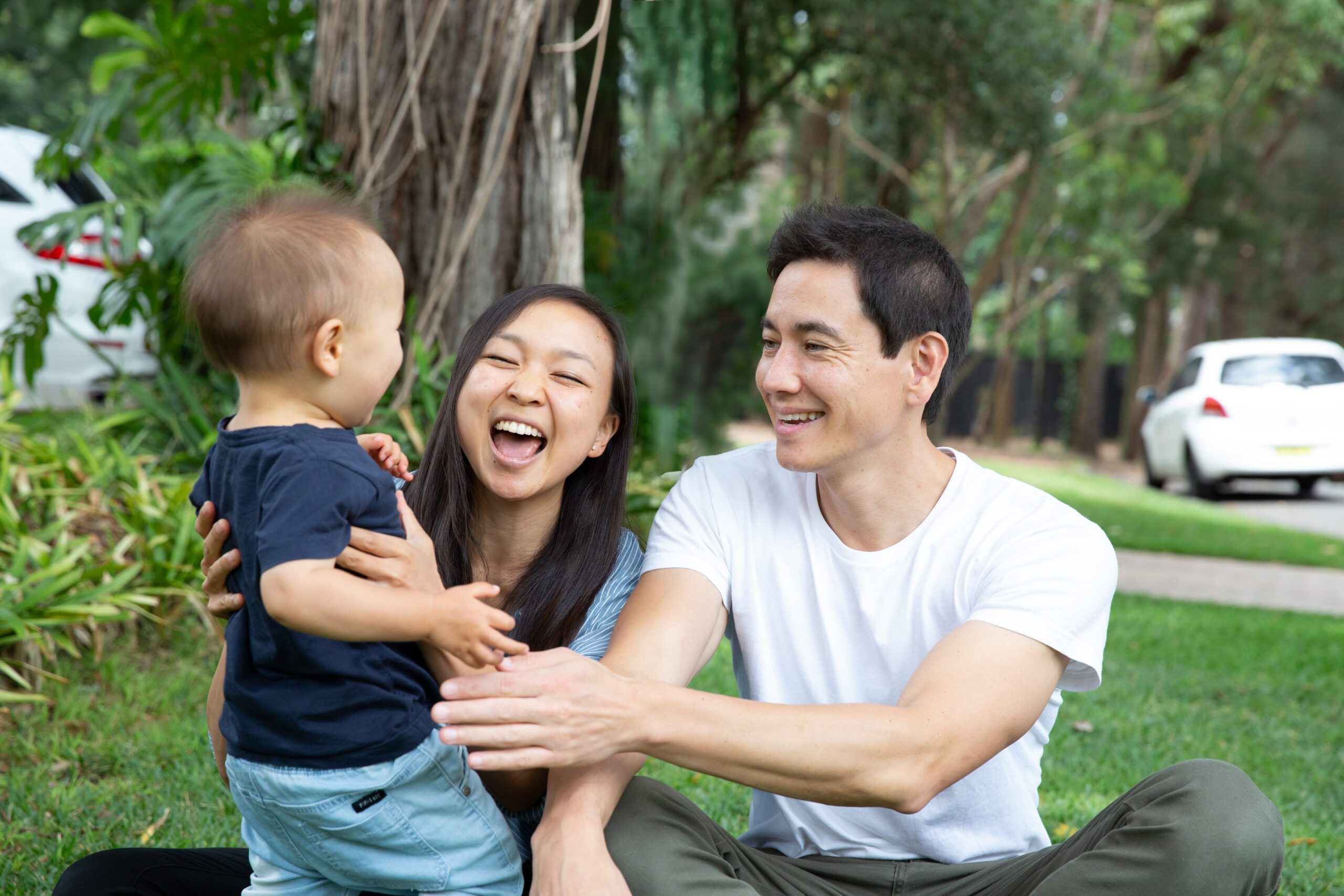 A young family with a toddler sit on grass and have a laugh.