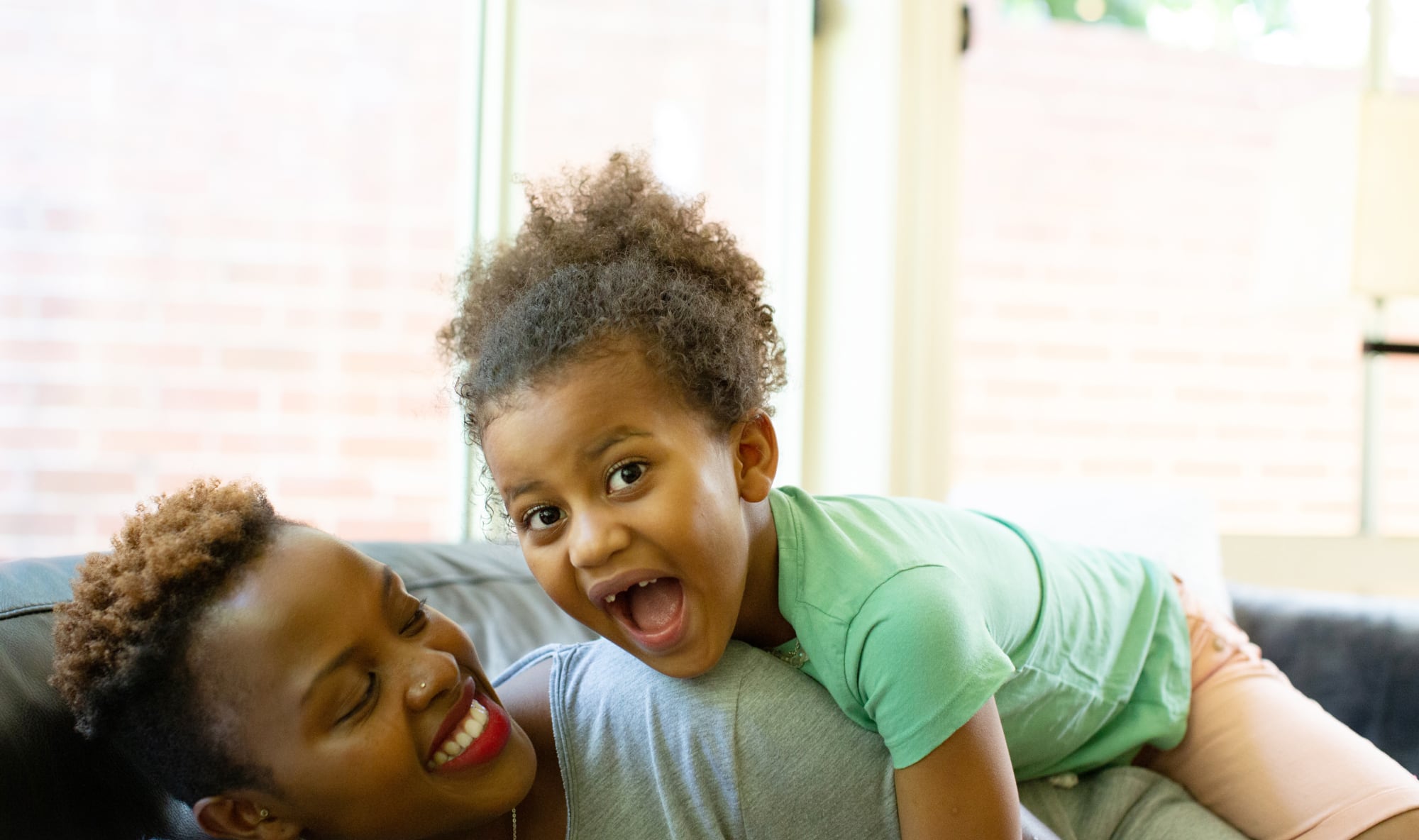 A mother and young child playing on the couch. The mother the child on top of her and the child has a big smile on their face.