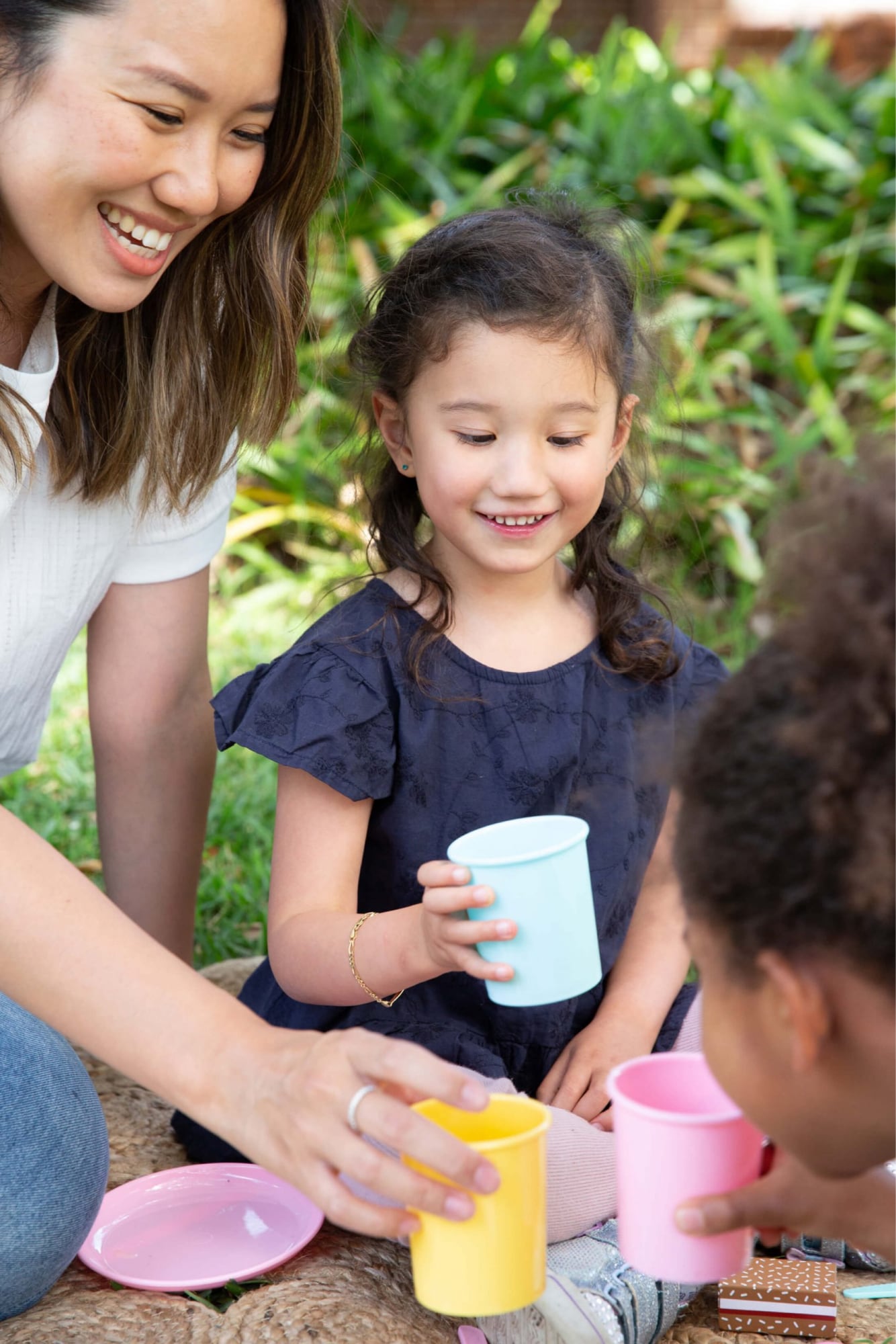 Two young children and a mother are playing with a tea set outside. The children and mother are smiling.