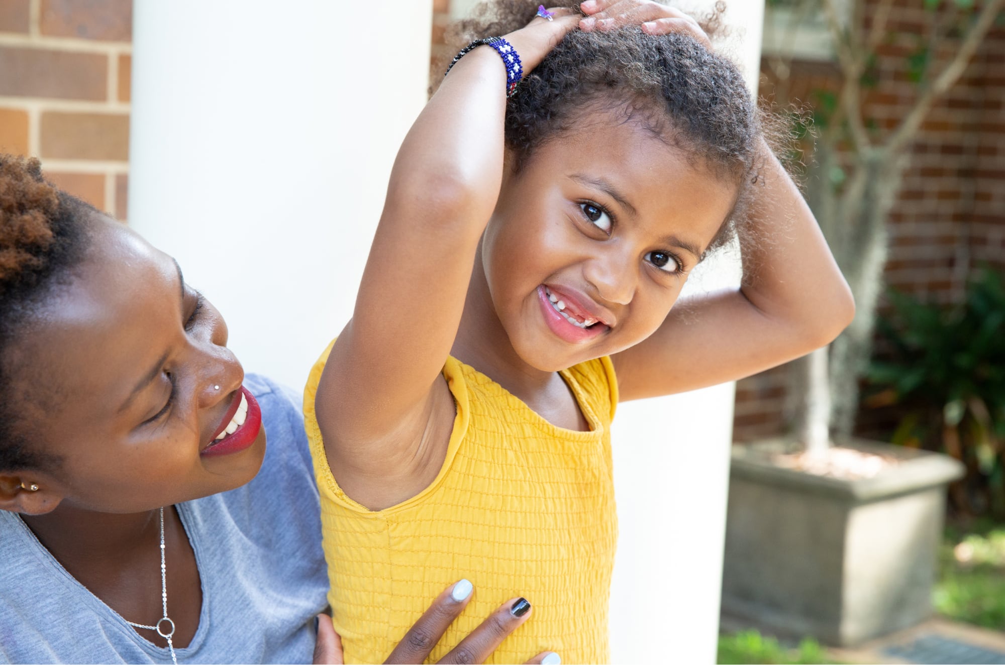 A young child smiling at the camera while their mother looks at them from behind. The mother is smiling at their child.