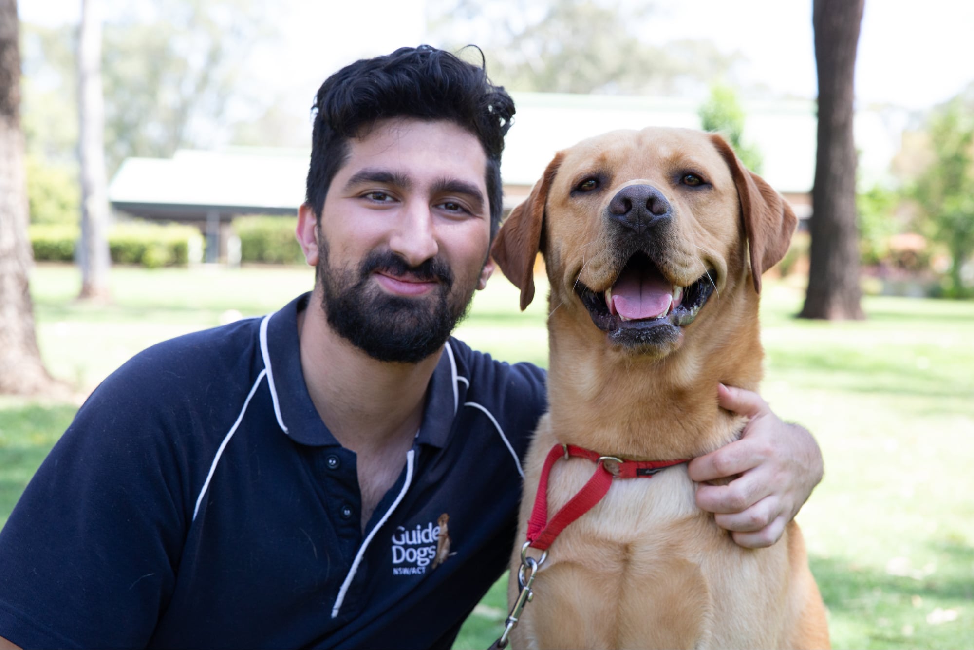A Guide Dogs staff member and a caramel labrador dog sitting outside. The staff member has their arm around the dog and both are looking at the camera smiling.