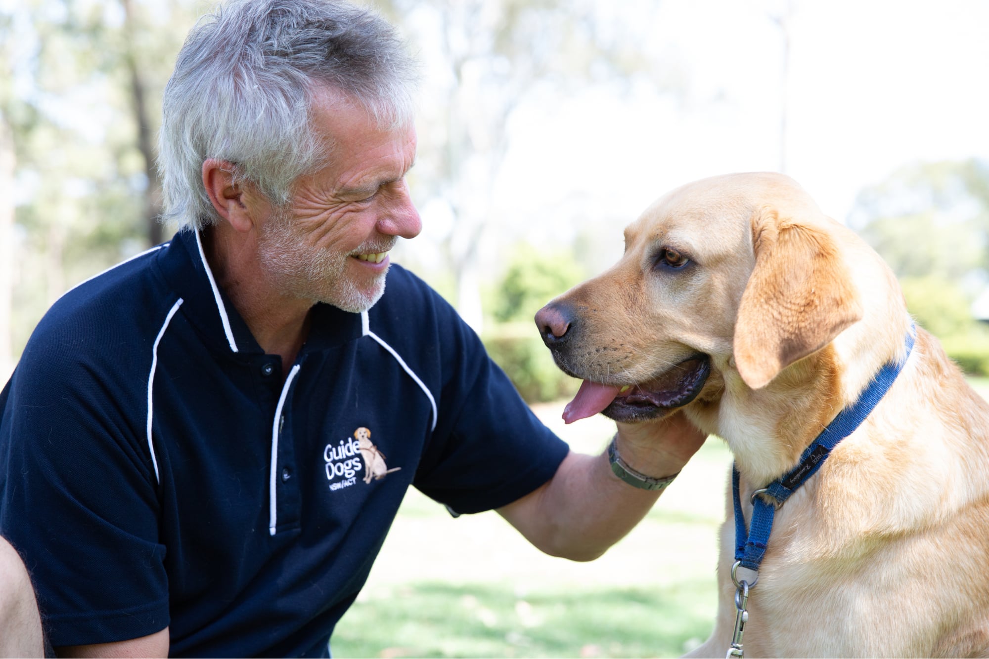 A Guide Dogs staff member and a yellow labrador dog sitting outside. The staff member is patting the dog whilst smiling and they are both looking at each other.