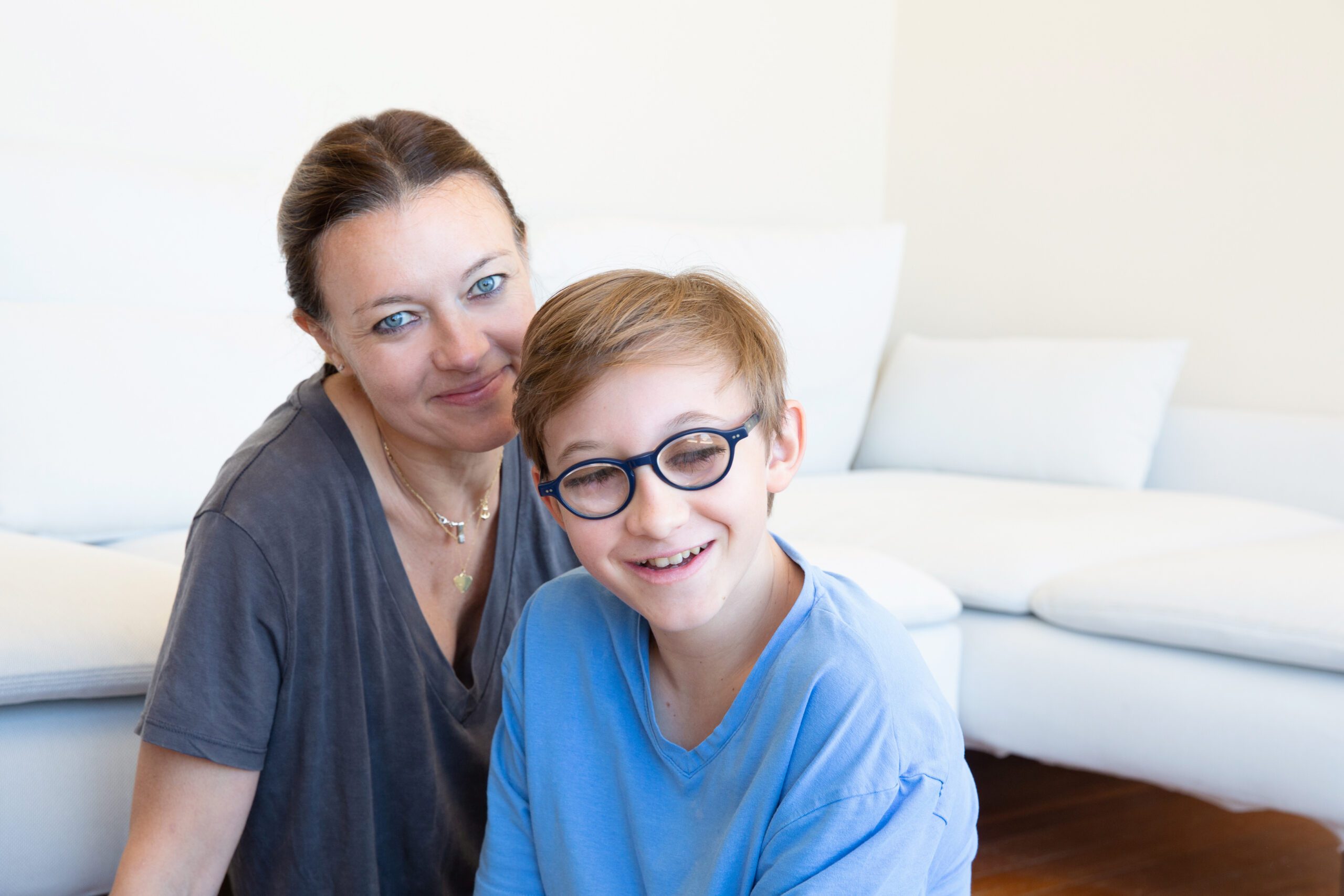 A young boy wearing a blue t-shirt and glasses sitting with his mum.