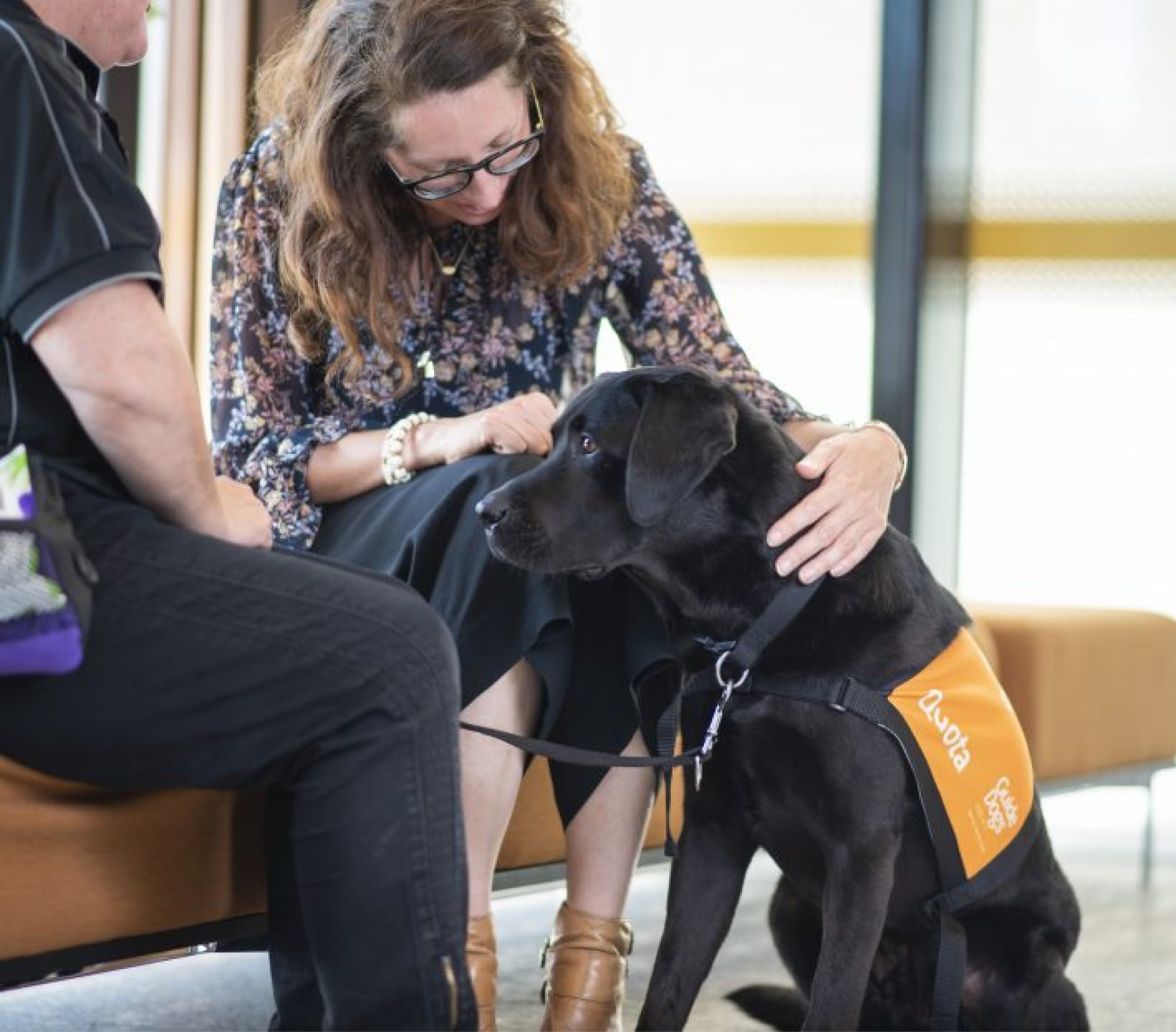 A person seated on a couch with a black labrador dog seated in front of the person. The person is looking down at the dog.