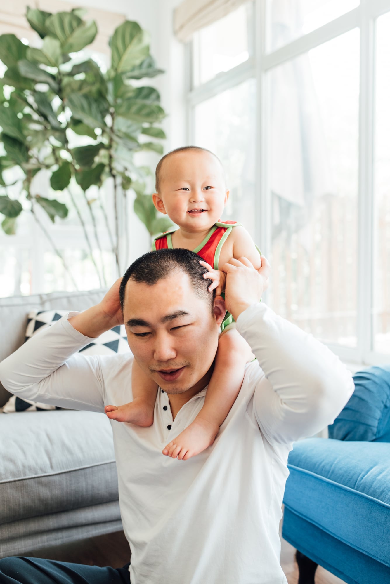 A young Asian blind man with his son at home. His son sits on his shoulders as he holds him around the waist. Big windows and greenery is in the background.