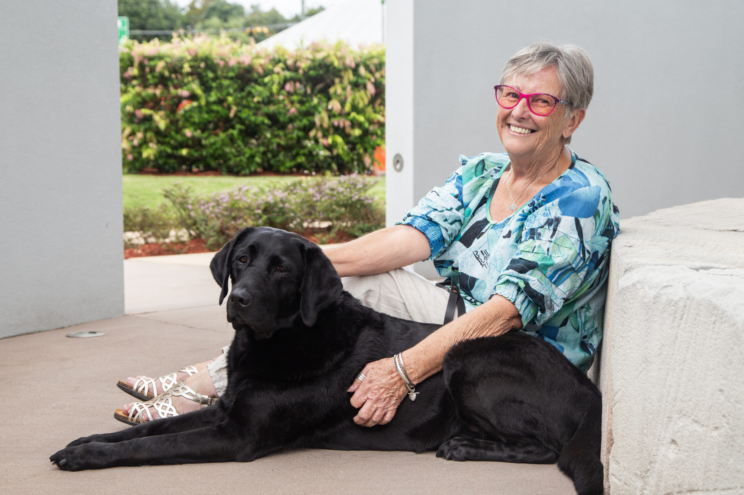 Guide Dogs Client, Sue sits on the concrete outside with her hand on black Guide Dog, Vegas.