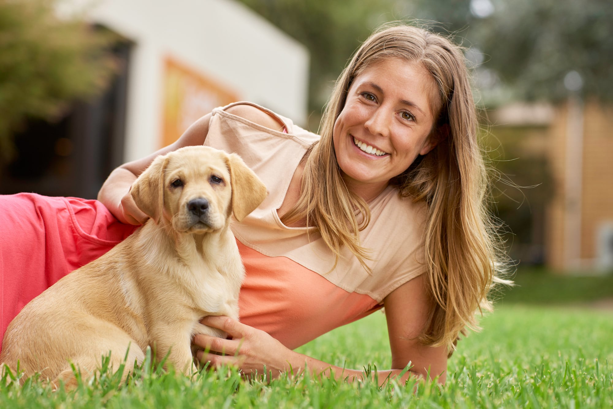 A person laying outside on some grass next to an eight week old labrador puppy. The puppy and person are both looking at the camera.