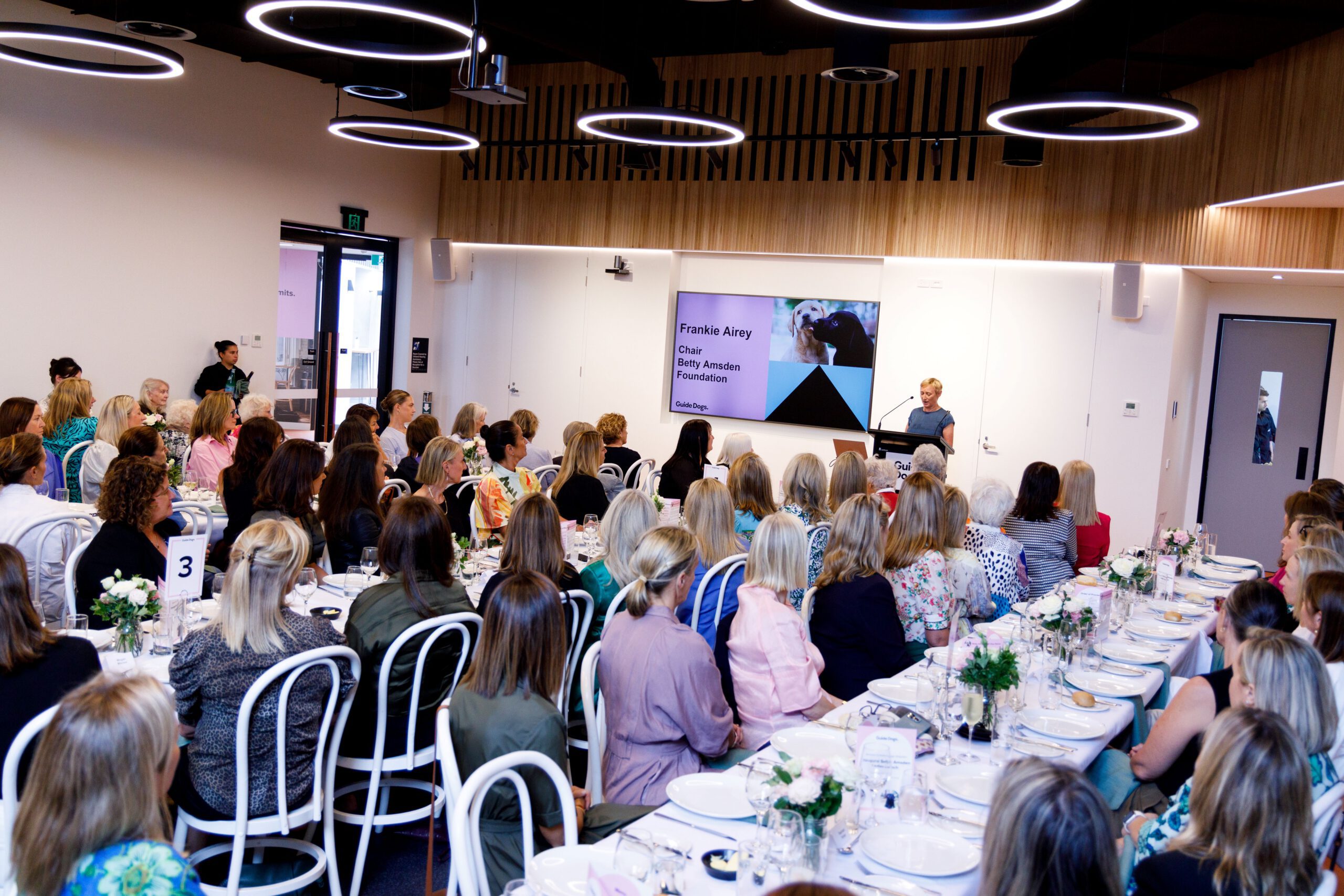 An image of people siting at three long tables, decorated with white tablecloths, flowers and cutlery. They are looking toward the front of the room, where someone is speaking at a lectern, and is located in front of a large digital screen.