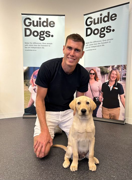 Mason Cox kneeling on the ground, with a yellow Labrador puppy next to him.