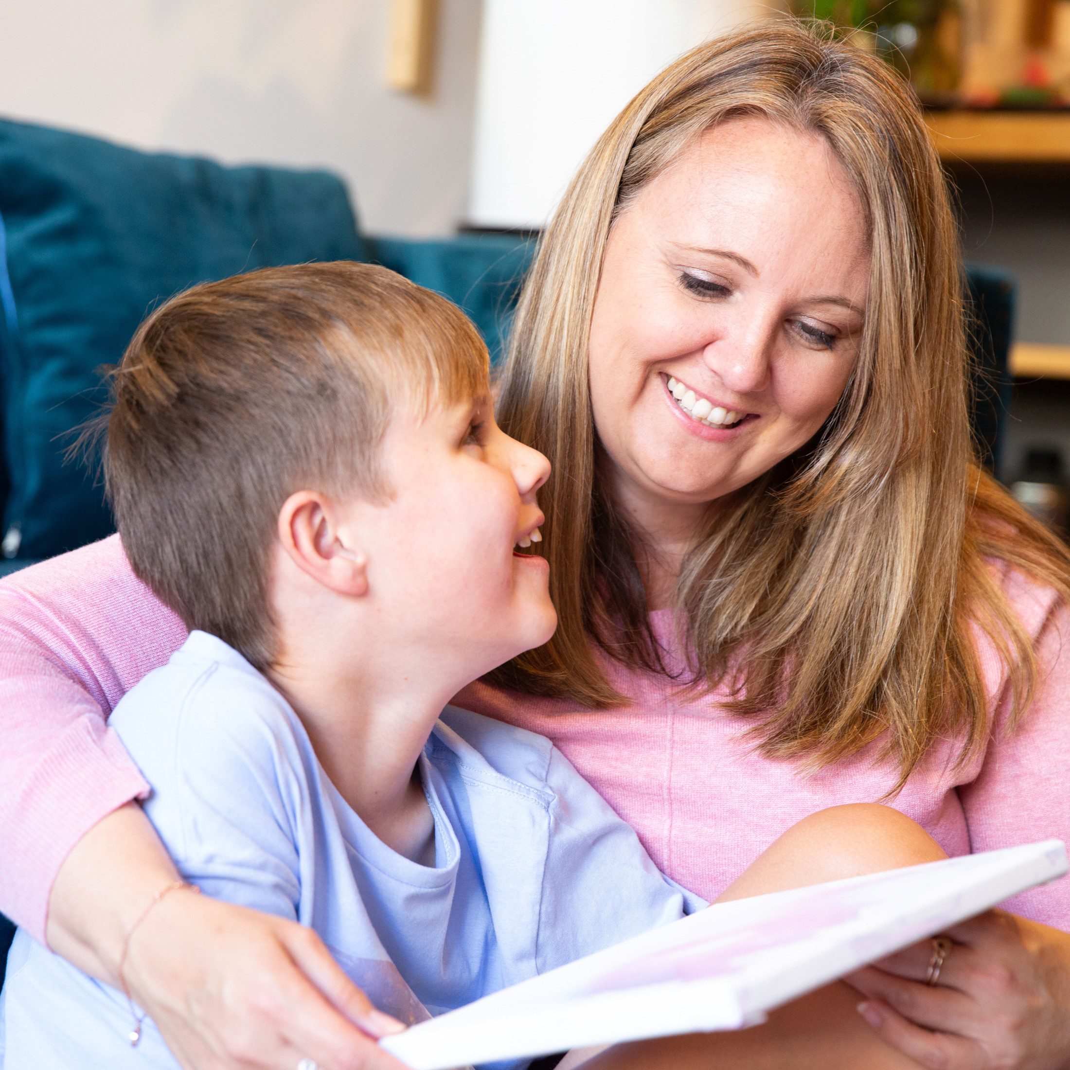 A parent with their arm wrapped around their child, holding a book. Seated in front of a couch. The parent and child are smiling at each other.