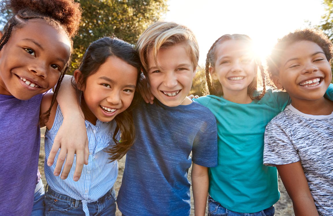 A group of 5 smiling kids are standing close together with their arms around each other smiling. They are outdoors and the sun is shining.