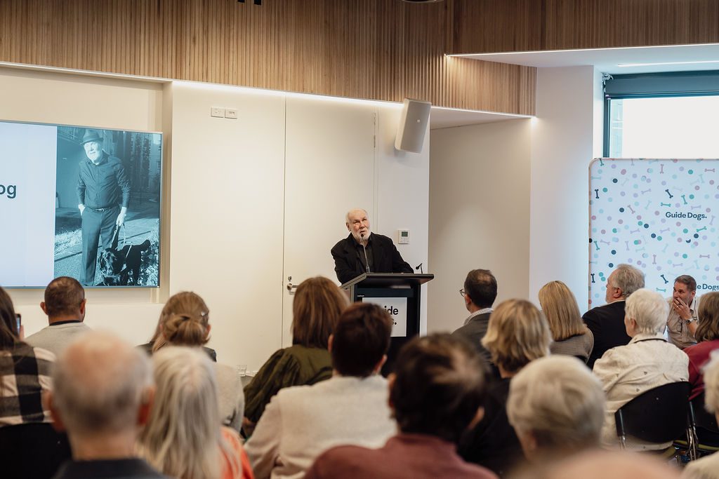 A person speaking at a lectern. A seated crowd facing the front of the room is listening to him speak.
