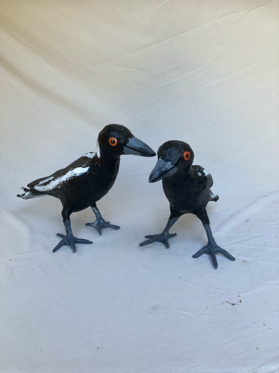 Two paper mache sculptures of black and white birds with orange eyes, standing on a coloured surface.