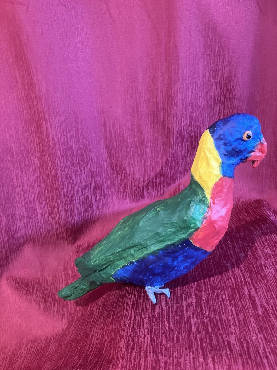 Colorful paper mache sculpture of a parrot with a blue head, yellow neck, green wings, red chest, and blue tail, set against a pink textured fabric background.