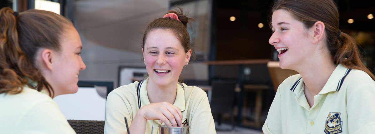 Three high school-aged students in school uniforms sit together at a café table, smiling and chatting over drinks. One student faces the camera while the other two are shown in profile, creating a relaxed and friendly atmosphere that reflects connection, confidence and social inclusion.
