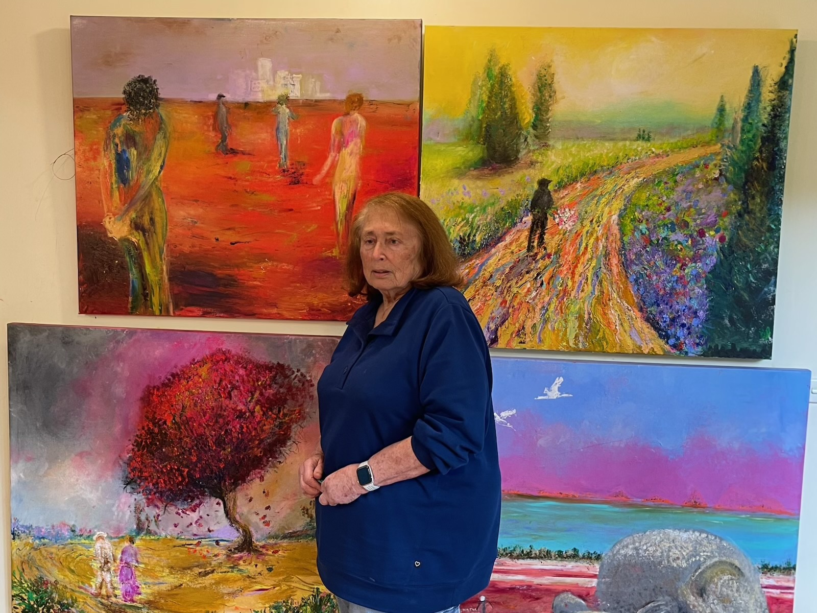 A woman stands in front of four colourful paintings displayed on a wall, including landscapes and figures. She wears a navy blue top and a smartwatch.