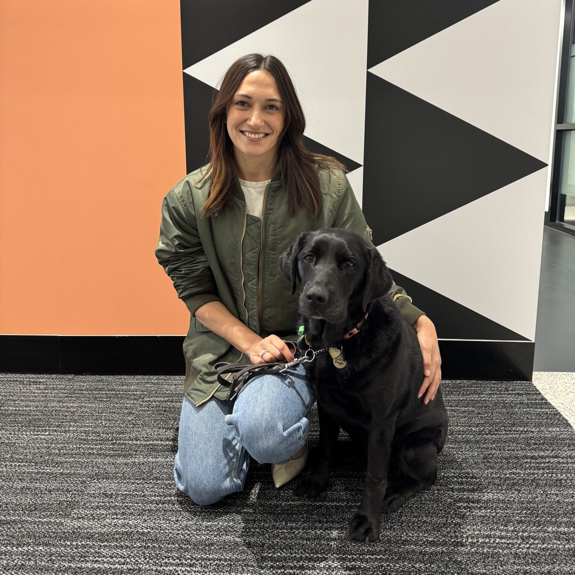 A smiling woman kneels beside a black Labrador wearing a harness and lead. She rests one hand on the dog’s back and holds its lead with the other. They are indoors in front of a black, white and orange geometric wall.
