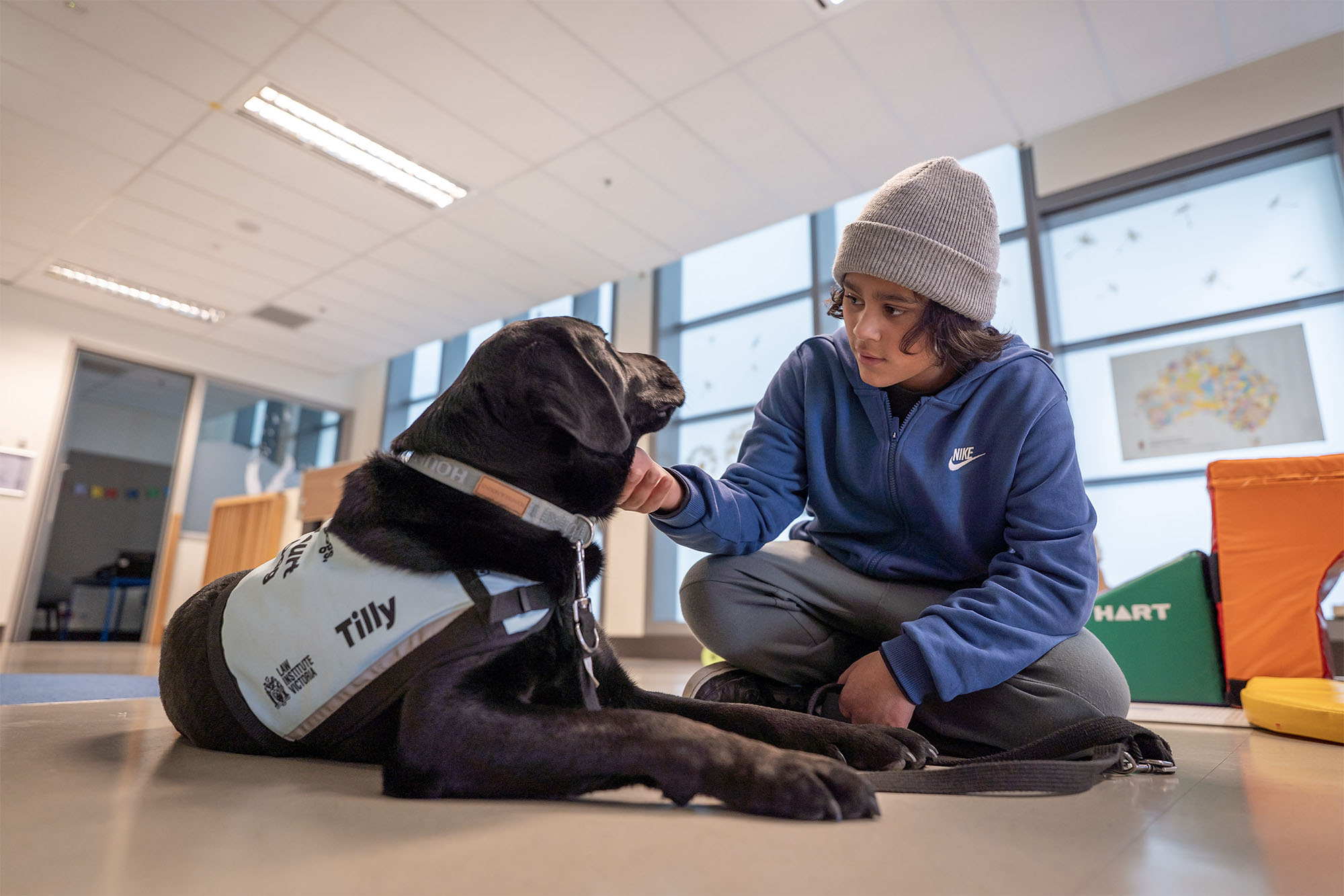 A young person sits on the floor in a classroom, gently patting Tilly, a black Labrador court dog wearing a light-coloured vest with her name displayed. Tilly is lying calmly on the floor, facing the young person. Colourful soft play equipment is visible in the background.