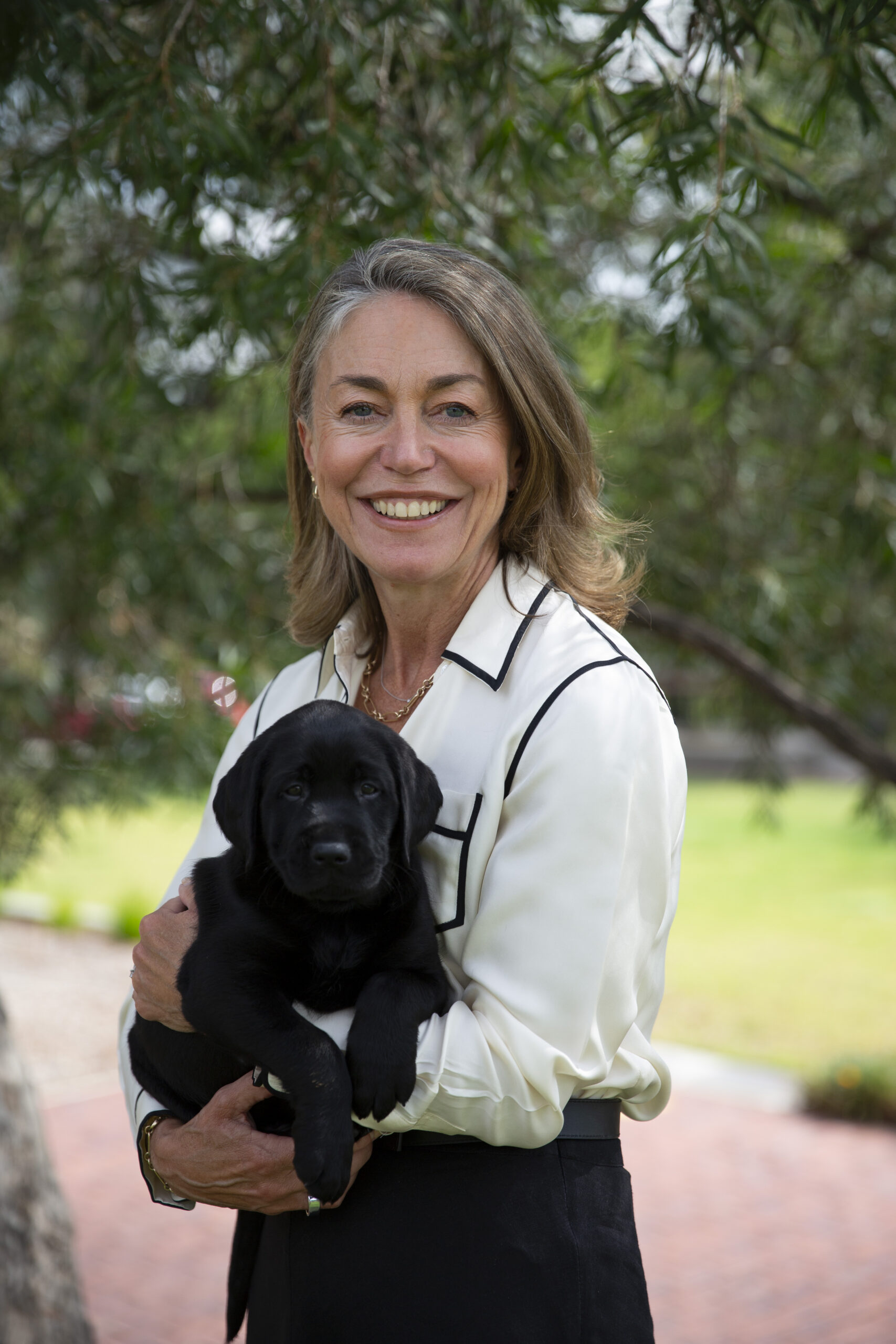 A smiling woman with shoulder-length brown hair stands outdoors holding a black Labrador puppy in her arms. She is wearing a white blouse with black trim and black trousers. Trees and greenery create a softly blurred background.