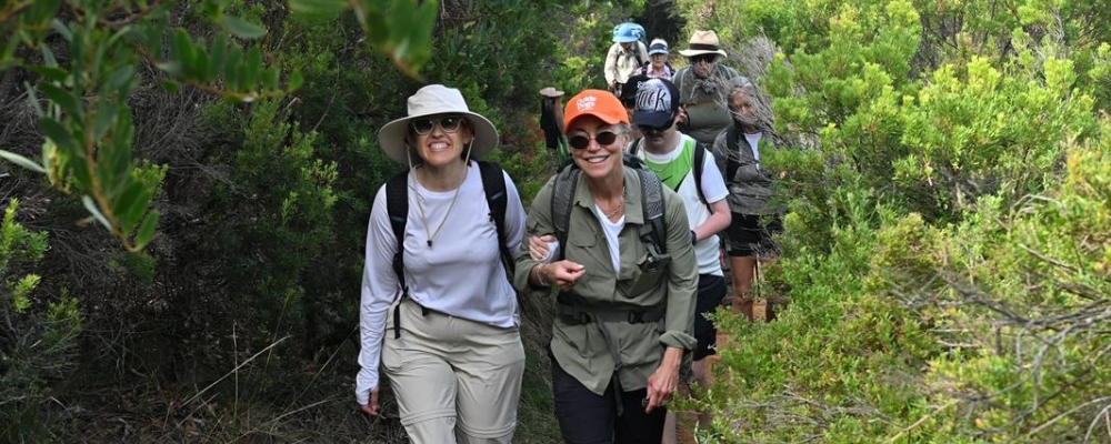 A group of hikers walking along a narrow bush track surrounded by dense green vegetation. Two women lead the group, smiling at the camera and wearing sun hats, sunglasses and backpacks. Several other hikers follow behind them along the trail.