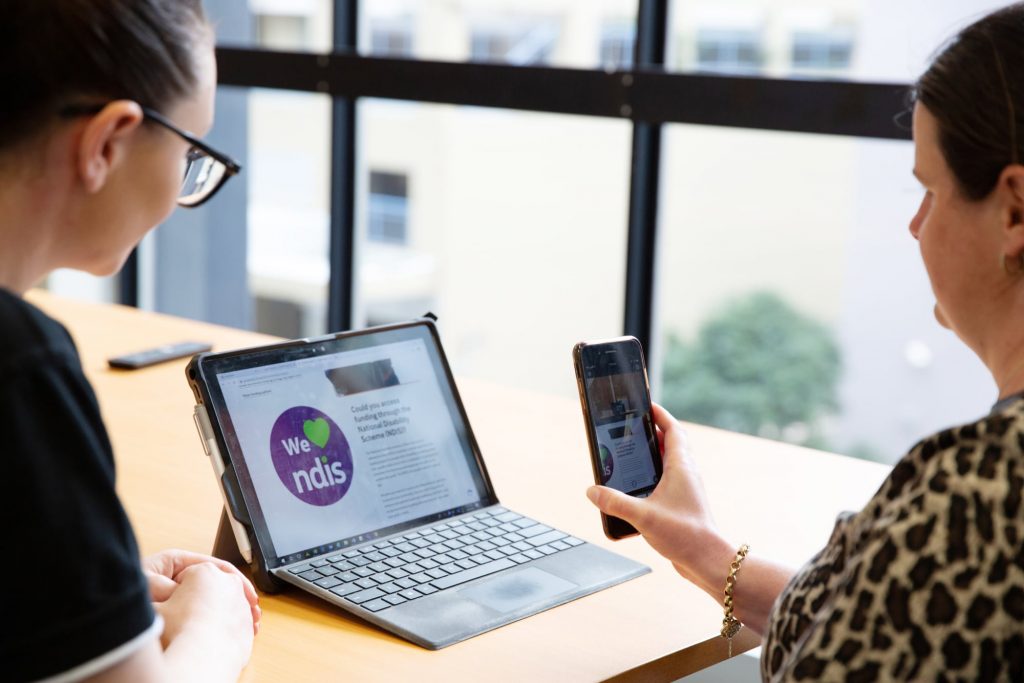 Two people looking at a laptop screen. One is holding a phone to the laptop screen and the other is watching on.