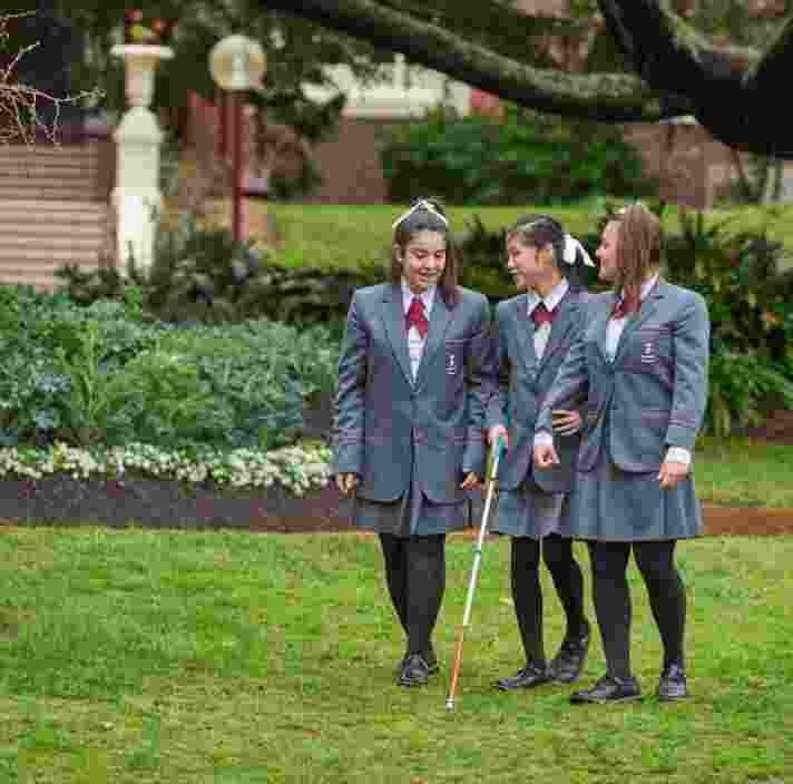 Three girls engaged in conversation as they walk through school grounds. The girl in the middle is using a cane.