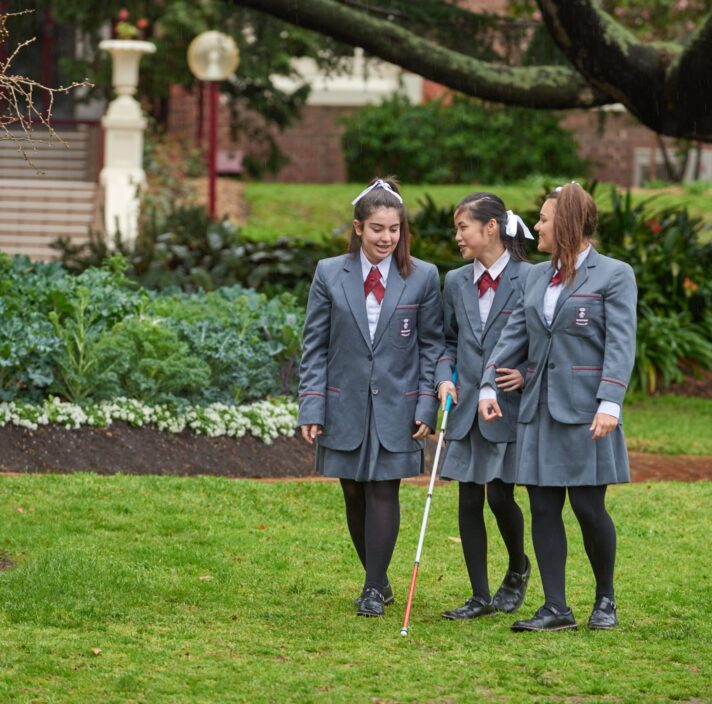 Three girls engaged in conversation as they walk through school grounds. The girl in the middle is using a cane.