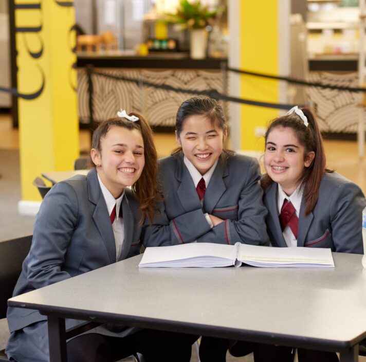 Three girls sitting at the school cafeteria, smiling.