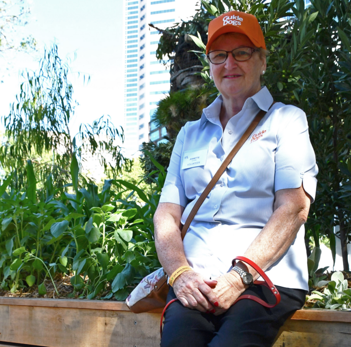 Puppy Raiser Jeanette sitting by a garden bed and wearing a Guide Dogs hat