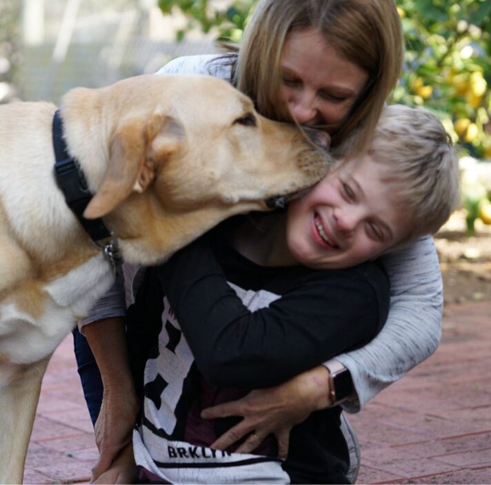 A young child is getting a kiss from a yellow labrador dog. His mother is behind them giving them a cuddle.
