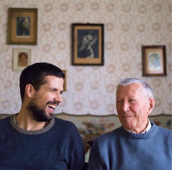 Two people, one adult & one older person, sitting on a couch looking at each other laughing.