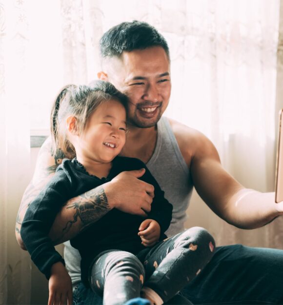 A young child sitting on their father's lap while they both smile while looking at an Ipad. The father has one had around his child and is holding the Ipad with the other hand.