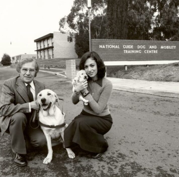 A 1940's black and white image of two people outside a Guide Dog Training Centre. One of the people in kneeling down next to a yellow labrador Guide Dog in harness and the other is holding a yellow ten week old labrador puppy. Both dogs and people are looking at the camera.