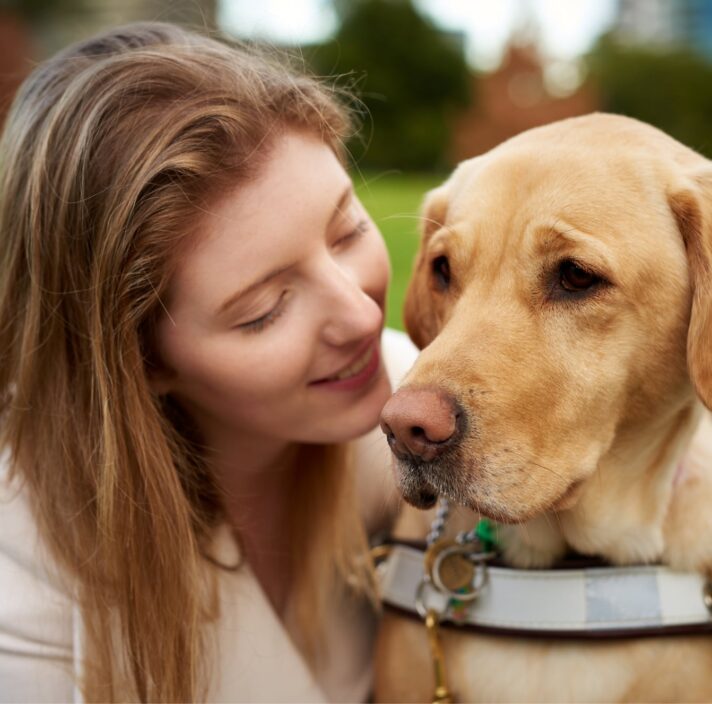 Close up of a person with their Guide Dog, who is caramel in colour and in harness. The person is smiling at the dog.