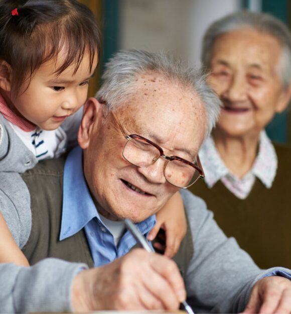 An older man writing while a young girl puts her arms around his shoulders, hugging him. The man is smiling and there is an older woman in the background who is not in focus but is also smiling.