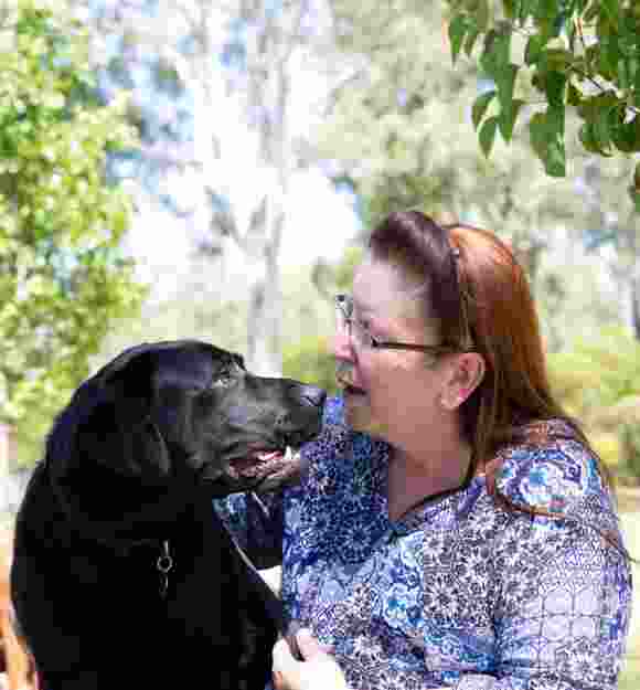 A person sitting outside with a black labrador dog. The person is looking at the dog smiling and the dog is looking at the person.
