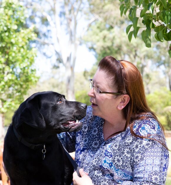 A person sitting outside with a black labrador dog. The person is looking at the dog smiling and the dog is looking at the person.
