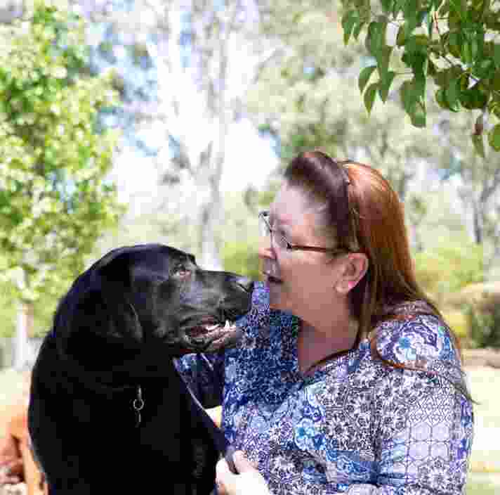 A person sitting outside with a black labrador dog. The person is looking at the dog smiling and the dog is looking at the person.