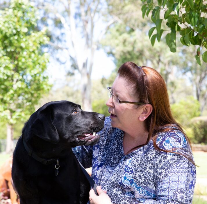 A person sitting outside with a black labrador dog. The person is looking at the dog smiling and the dog is looking at the person.