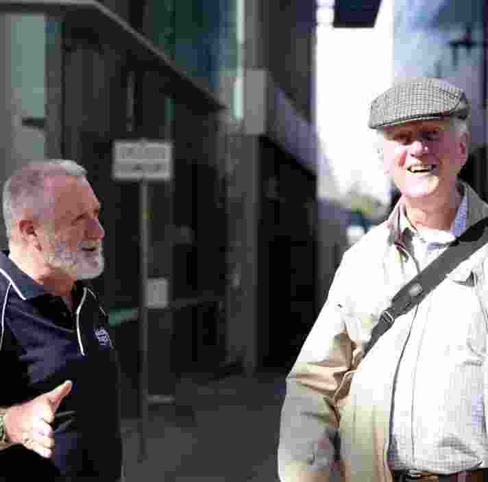 A Guide Dogs staff members is talking to an older adult in the street. The an older adult is looking at the camera with a smile on his face.